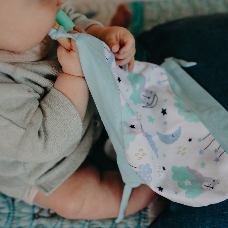 Introducing Cuddle Soother: The perfect blend of comfort and teething relief. Sleepy Bear design laid flat on a plain white background.. Baby holding blanket and chewing on teether.