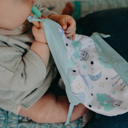 Introducing Cuddle Soother: The perfect blend of comfort and teething relief. Sleepy Bear design laid flat on a plain white background.. Baby holding blanket and chewing on teether.